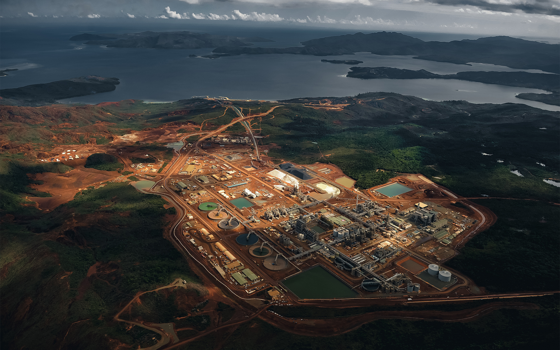 Aerial shot of a large industrial facility near a coastline surrounded by forest and mountains.